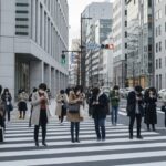 2026年5月時点の自動車保険一括見積もりサービス利用時の注意点について、都市の風景と携帯電話を使う人々を描いた日常シーンの画像。