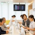 「紙の保険証が終了」クリニックでの診察の様子。患者とスタッフが健康保険の変更への案内を受けるシーンを描写し、理解を求める雰囲気を伝えます。