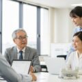 再雇用時の社会保険に関するガイド。オフィスでの仕事をするシニアと若手社員の交流を描写した画像。