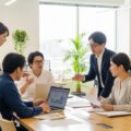 2026年の社会保険料、106万円・130万円の壁に関するダブル改正の動向を示す現代の職場風景。多様な人々が議論しているシーン。